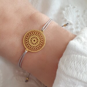 Puede incluir: Un brazalete dorado con un delicado colgante circular que presenta un dise&ntilde;o de mandala detallado. El brazalete est&aacute; hecho con un cord&oacute;n gris y es ajustable para un ajuste c&oacute;modo.
