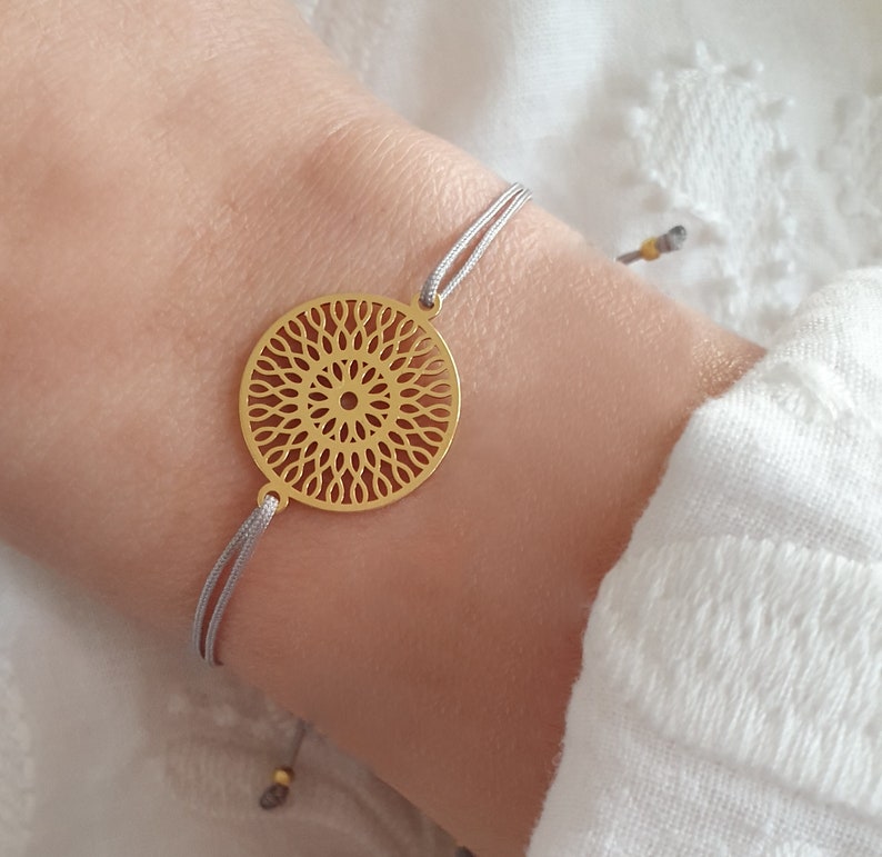 Puede incluir: Un brazalete dorado con un delicado colgante circular que presenta un dise&ntilde;o de mandala detallado. El brazalete est&aacute; hecho con un cord&oacute;n gris y es ajustable para un ajuste c&oacute;modo.