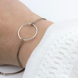 Könnte beinhalten: Ein schlichtes Armband mit einem silbernen Kreisanhänger und einer braunen Schnur. Die Schnur ist mit einem Schiebeknoten verstellbar.