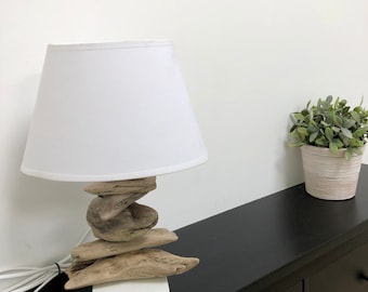 May include: A white lampshade with a white base sits on a black surface. The lamp base is made of stacked pieces of driftwood.