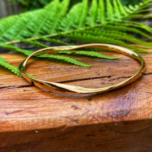 May include: A polished gold-tone bangle bracelet featuring a continuous, smooth, and undulating twisted design. The circular metal band has a reflective, high-shine finish that catches the light, creating subtle highlights along its curved surface.