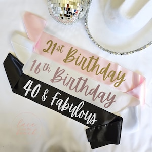 May include: Three satin sashes in pink, white and black with gold and pink lettering. The sashes read "21st Birthday", "16th Birthday", and "40 & Fabulous".