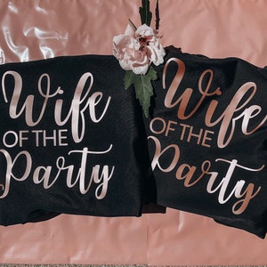 Op de afbeelding: Twee zwarte T-shirts met de tekst "Wife of the Party" in ros&eacute;gouden letters.