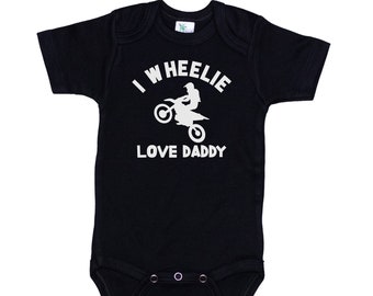 May include: Black baby onesie with white text that reads "I Wheelie Love Daddy" and a white silhouette of a person riding a dirt bike.