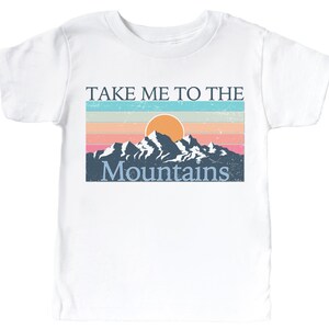May include: White t-shirt with a vintage-style graphic of a mountain range with a setting sun and the text "Take Me To The Mountains".