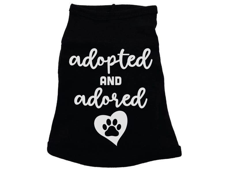 May include: Black dog shirt with white text that says "adopted and adored" with a white paw print inside a heart.