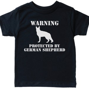 Pu&ograve; includere: Una maglietta nera a maniche corte con testo bianco che recita "WARNING PROTECTED BY GERMAN SHEPHERD". Sopra il testo &egrave; presente una silhouette bianca di un pastore tedesco.