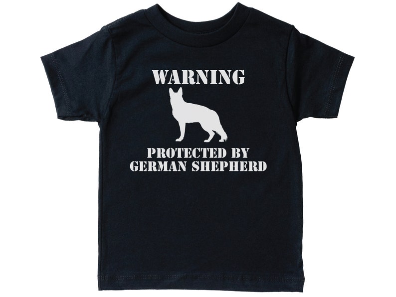 Pu&ograve; includere: Una maglietta nera a maniche corte con testo bianco che recita "WARNING PROTECTED BY GERMAN SHEPHERD". Sopra il testo &egrave; presente una silhouette bianca di un pastore tedesco.