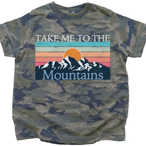 May include: A camouflage green t-shirt with a vintage-style graphic of mountains and a sunset. The text on the shirt reads "Take Me To The Mountains".
