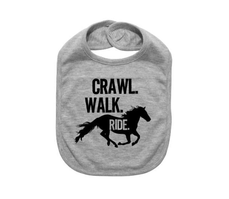 May include: A gray baby bib with the words "Crawl. Walk. Ride." in black block letters. A black silhouette of a running horse is below the text. The bib has a rounded shape and a snap closure.