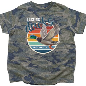 May include: A camouflage t-shirt with a retro-style graphic of a duck flying in front of a sunset. The text on the shirt reads "I like big ducks and I cannot lie."
