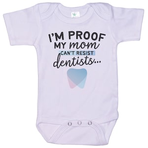 May include: A lavender baby onesie with short sleeves and a white graphic that says "I'm proof my mom can't resist dentists..." with a blue and pink tooth graphic.