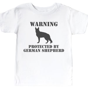Pu&ograve; includere: Maglietta bianca con una silhouette nera di un pastore tedesco e il testo "WARNING PROTECTED BY GERMAN SHEPHERD".