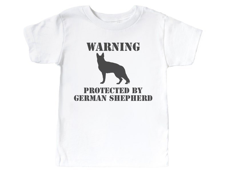 Pu&ograve; includere: Maglietta bianca con una silhouette nera di un pastore tedesco e il testo "WARNING PROTECTED BY GERMAN SHEPHERD".