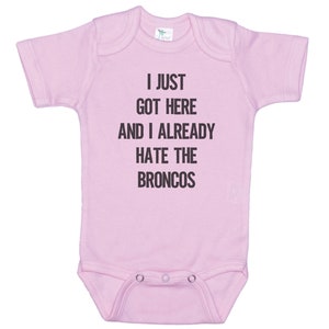 May include: Pink baby onesie with the text "I just got here and I already hate the Broncos" printed in black.