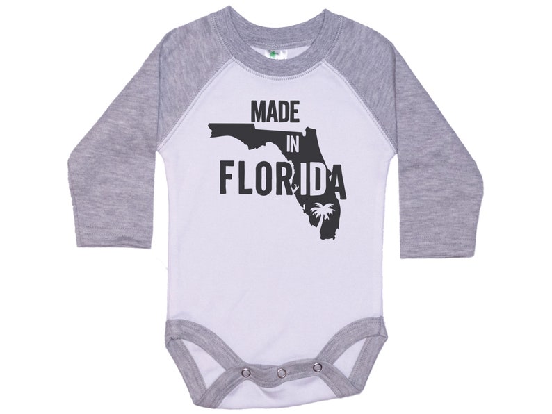 K&ouml;nnte beinhalten: Wei&szlig;er und grauer Langarm-Babybody mit einer schwarzen Umrandung des Bundesstaates Florida und dem Text "MADE IN FLORIDA".