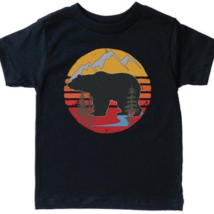 Może przedstawiać: Czarny T-shirt z grafiką w stylu retro przedstawiającą niedźwiedzia stojącego przed górami i zachodem słońca.