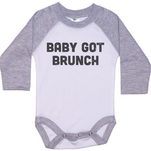 May include: A white and grey long-sleeved baby onesie with a baseball style design. The onesie has the text "BABY GOT BRUNCH" printed in black.