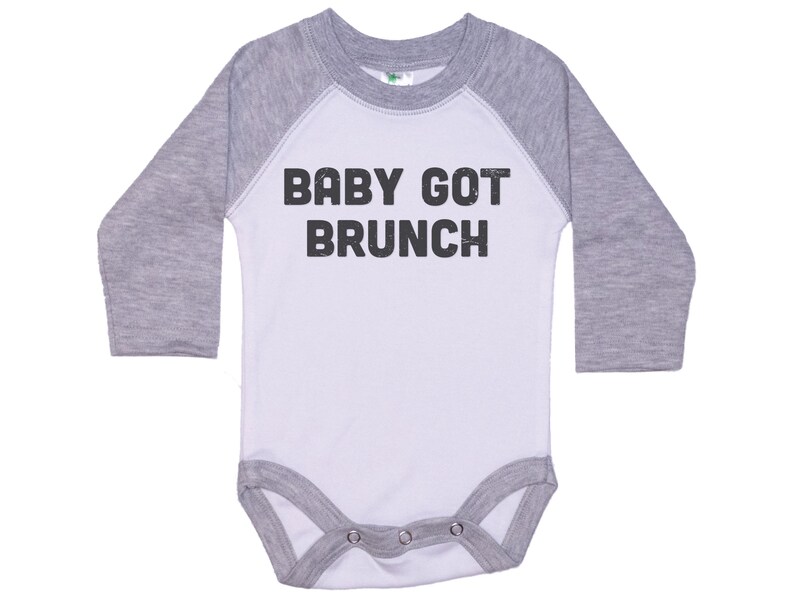 May include: A white and grey long-sleeved baby onesie with a baseball style design. The onesie has the text "BABY GOT BRUNCH" printed in black.