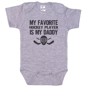May include: Gray baby onesie with black text that reads "My favorite hockey player is my daddy" with a hockey player's mask and two hockey sticks.