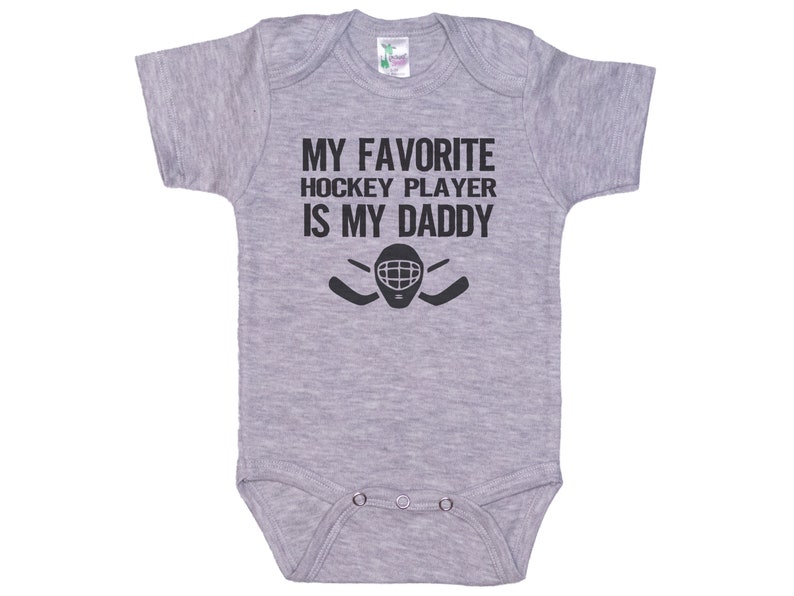 May include: Gray baby onesie with black text that reads "My favorite hockey player is my daddy" with a hockey player's mask and two hockey sticks.