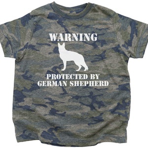 Pu&ograve; includere: Una maglietta verde mimetica con il testo "WARNING PROTECTED BY GERMAN SHEPHERD" e una silhouette bianca di un pastore tedesco.