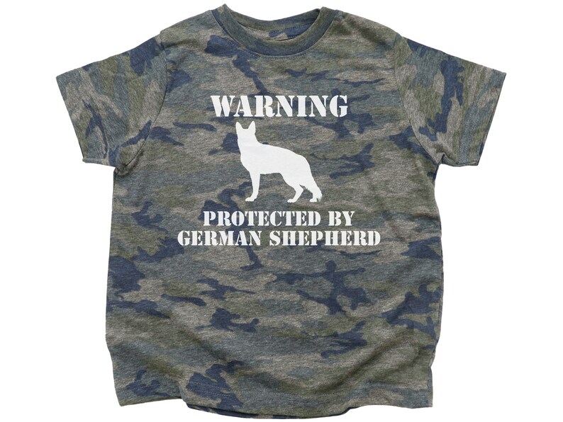 Pu&ograve; includere: Una maglietta verde mimetica con il testo "WARNING PROTECTED BY GERMAN SHEPHERD" e una silhouette bianca di un pastore tedesco.