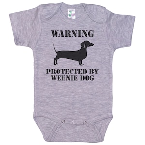 May include: Gray baby onesie with a black silhouette of a dachshund and the text "WARNING PROTECTED BY WEENIE DOG"