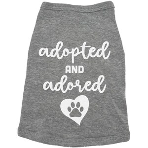 May include: Gray dog shirt with white text that says "adopted and adored" with a white paw print inside a heart.