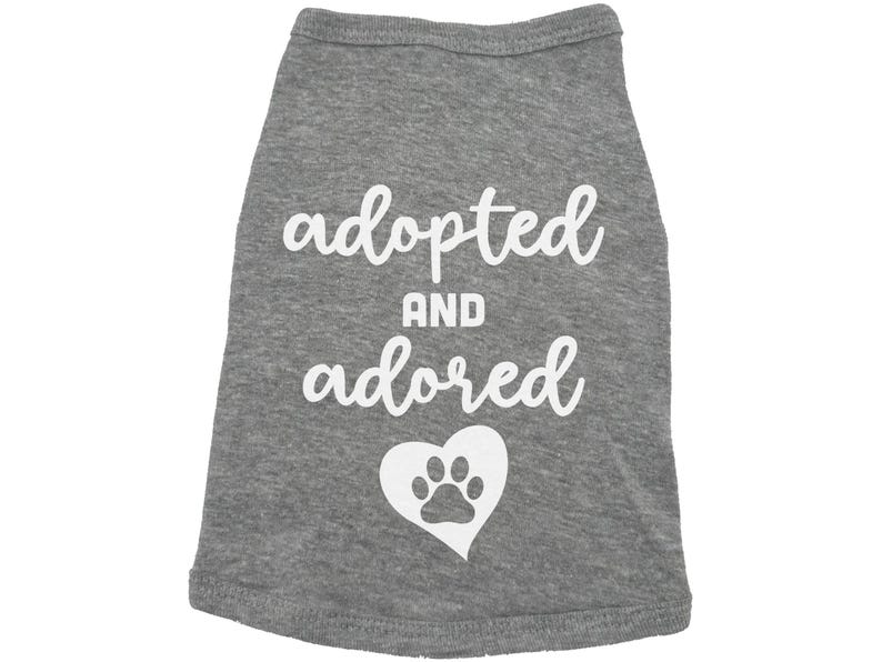 May include: Gray dog shirt with white text that says "adopted and adored" with a white paw print inside a heart.