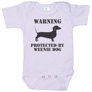 May include: White baby onesie with black text that reads "WARNING PROTECTED BY WEENIE DOG" and a black silhouette of a dachshund.