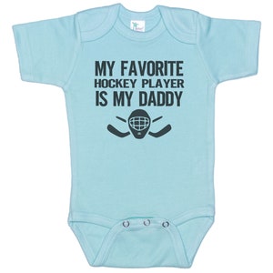 May include: Light blue baby onesie with a black graphic that says "My favorite hockey player is my daddy" with a hockey mask and sticks.