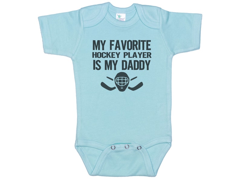 May include: Light blue baby onesie with a black graphic that says "My favorite hockey player is my daddy" with a hockey mask and sticks.