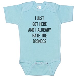 May include: A light blue baby onesie with the text "I just got here and I already hate the Broncos" printed in black.