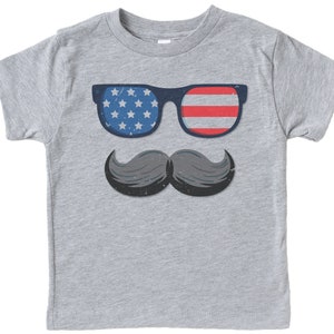 May include: A grey t-shirt with a graphic of sunglasses with a blue frame and the American flag design on the lenses, and a grey moustache. The sunglasses have a blue frame with white stars on one lens and red and white stripes on the other. The moustache is grey.