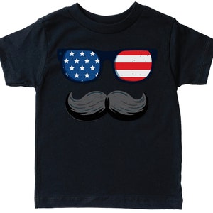 May include: A navy blue t-shirt with a graphic of sunglasses and a moustache. The sunglasses have a blue frame, with one lens showing white stars on a blue background and the other with red and white stripes, representing the American flag.