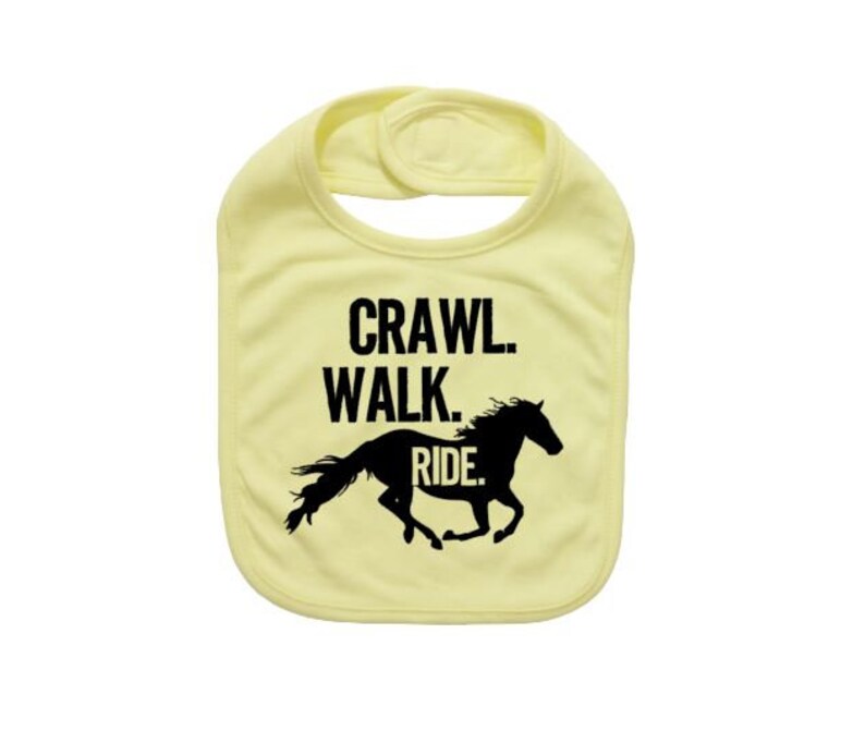 May include: A light yellow baby bib with the words "Crawl. Walk. Ride." in black block letters. A black silhouette of a running horse is below the text. The bib has a rounded shape and a snap closure.