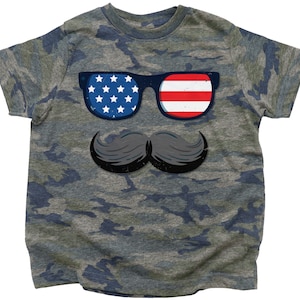 May include: A camouflage-patterned t-shirt with a graphic of sunglasses displaying the American flag and a black mustache. The sunglasses have a blue frame. The shirt is a youth size.