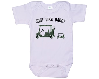 May include: White baby onesie with short sleeves and a green graphic of two golf carts. The text "JUST LIKE DADDY" is printed in black above the golf carts.