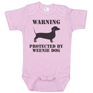May include: Pink baby onesie with a black dachshund silhouette and the text "WARNING PROTECTED BY WEENIE DOG".