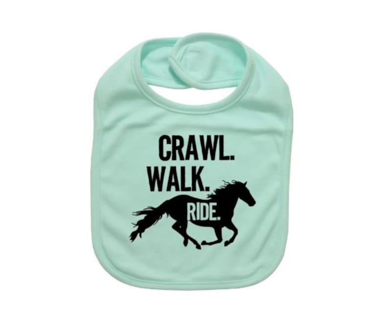 May include: A light green baby bib with the words "Crawl. Walk. Ride." in black block letters above a black silhouette of a running horse. The bib has a rounded shape and a snap closure at the neck.
