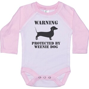 May include: A pink and white long-sleeved baby onesie with a black silhouette of a dachshund and the text "WARNING PROTECTED BY WEENIE DOG".