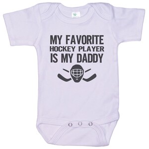May include: White baby onesie with black text that reads "My favorite hockey player is my daddy" with a hockey goalie mask and two hockey sticks.