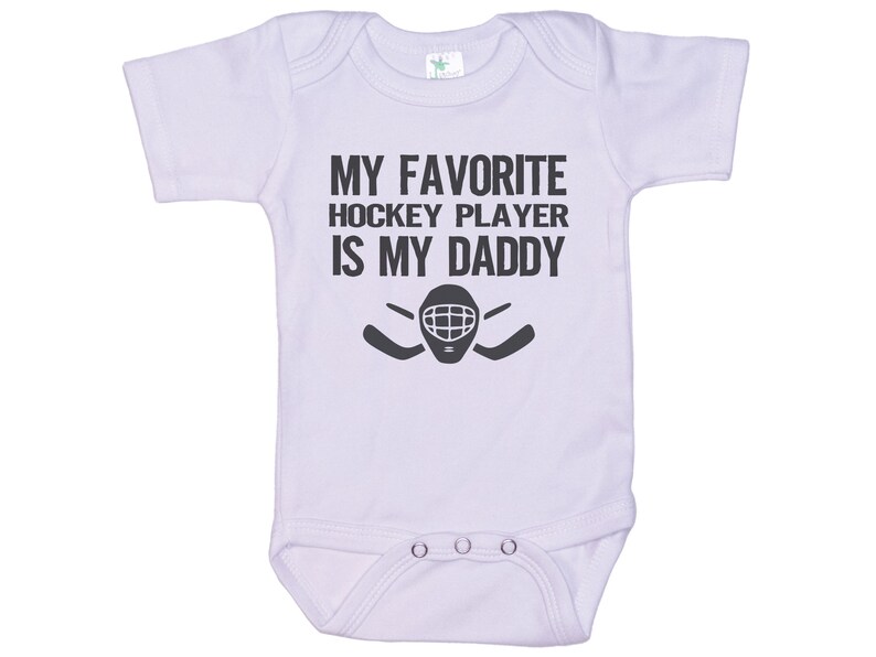 May include: White baby onesie with black text that reads "My favorite hockey player is my daddy" with a hockey goalie mask and two hockey sticks.