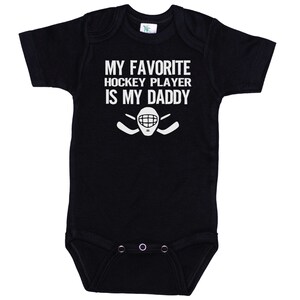May include: Black baby onesie with white text that reads "My favorite hockey player is my daddy" with a hockey stick and goalie mask graphic.