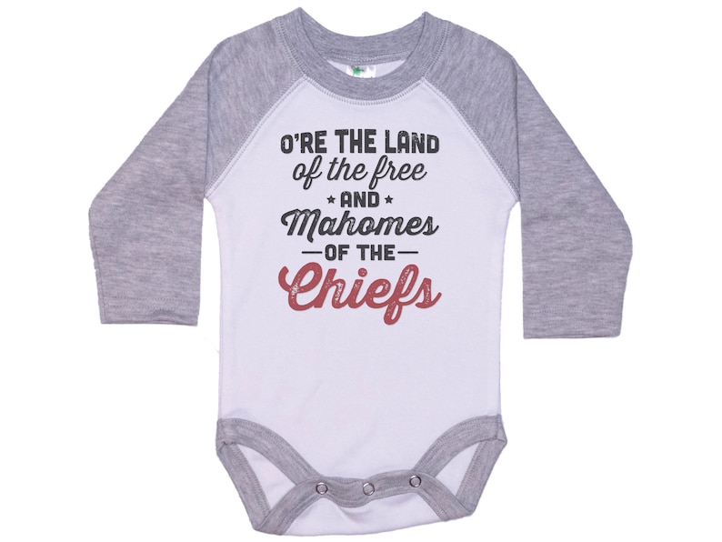 Pode incluir: Macac&atilde;o de beb&ecirc; de manga comprida branco e cinza com um design estilo beisebol. O macac&atilde;o tem o texto "O'RE THE LAND of the free * AND * Mahomes -OF THE- Chiefs" em preto e vermelho.