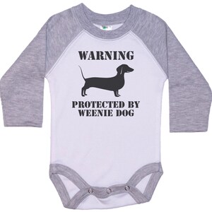 May include: A white and gray long-sleeve baby onesie with a black silhouette of a dachshund and the text "WARNING PROTECTED BY WEENIE DOG".