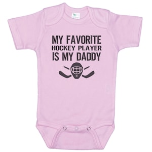 May include: Pink baby onesie with a black graphic that says "My favorite hockey player is my daddy" with a hockey mask and sticks.