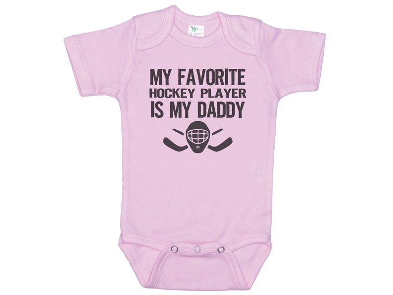 May include: Pink baby onesie with a black graphic that says "My favorite hockey player is my daddy" with a hockey mask and sticks.