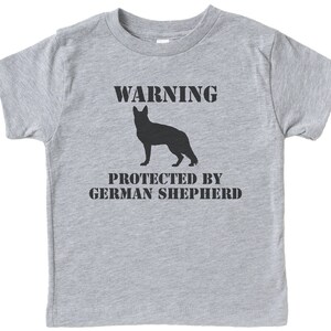 Pu&ograve; includere: Maglietta grigia a maniche corte con una silhouette nera di un pastore tedesco e il testo "WARNING PROTECTED BY GERMAN SHEPHERD".
